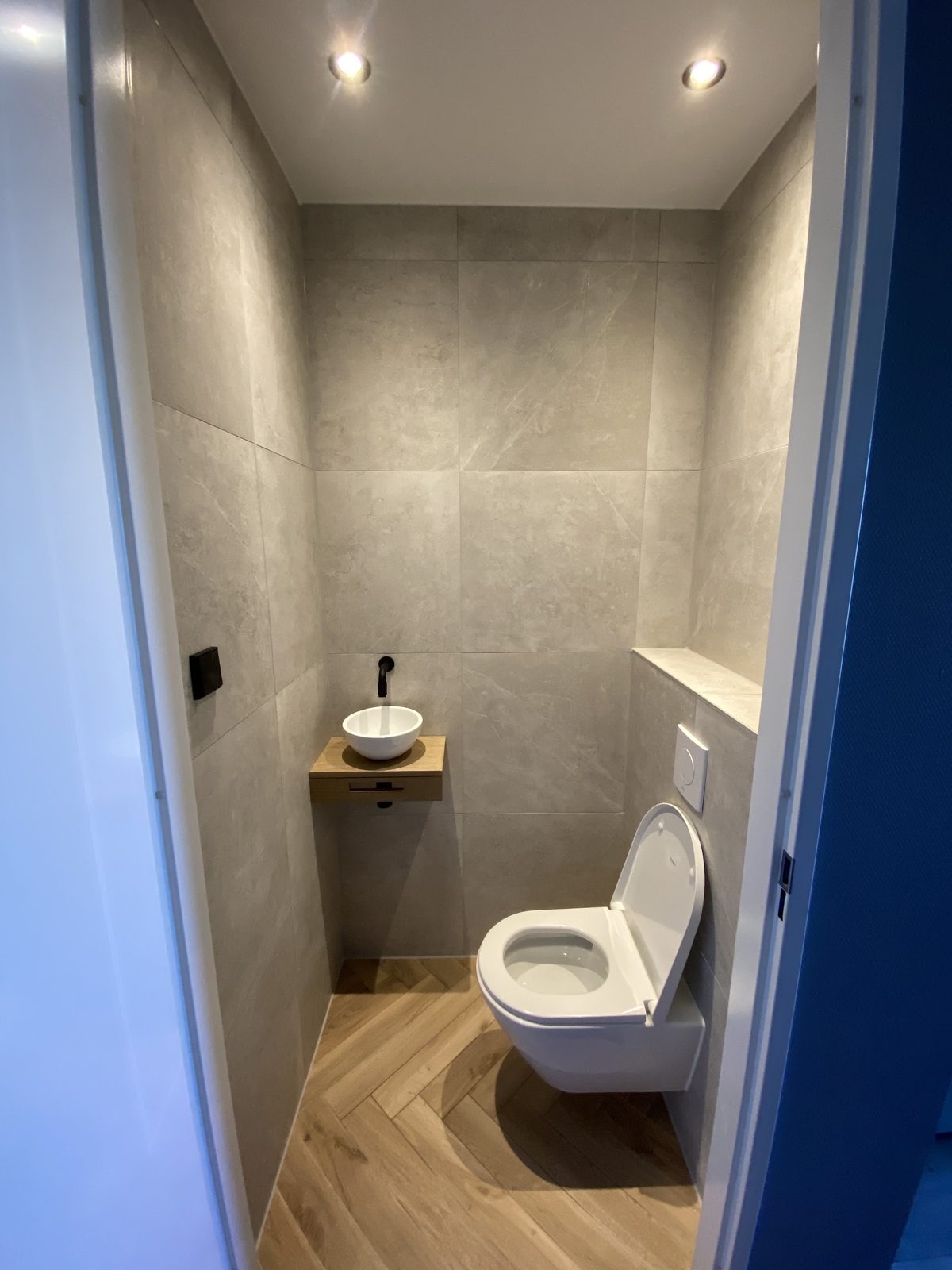 Toilet met grote grijze wandtegels, visgraat houtlook vloer en een kleine waskom op een houten plankje
