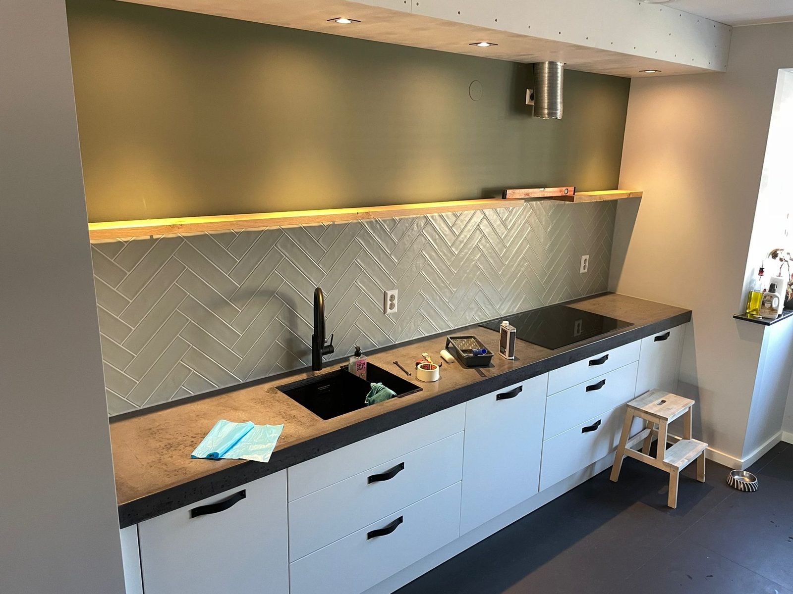 Keuken met groengrijze visgraat-tegelwand achter het aanrecht, witte kasten en houten plank met verlichting