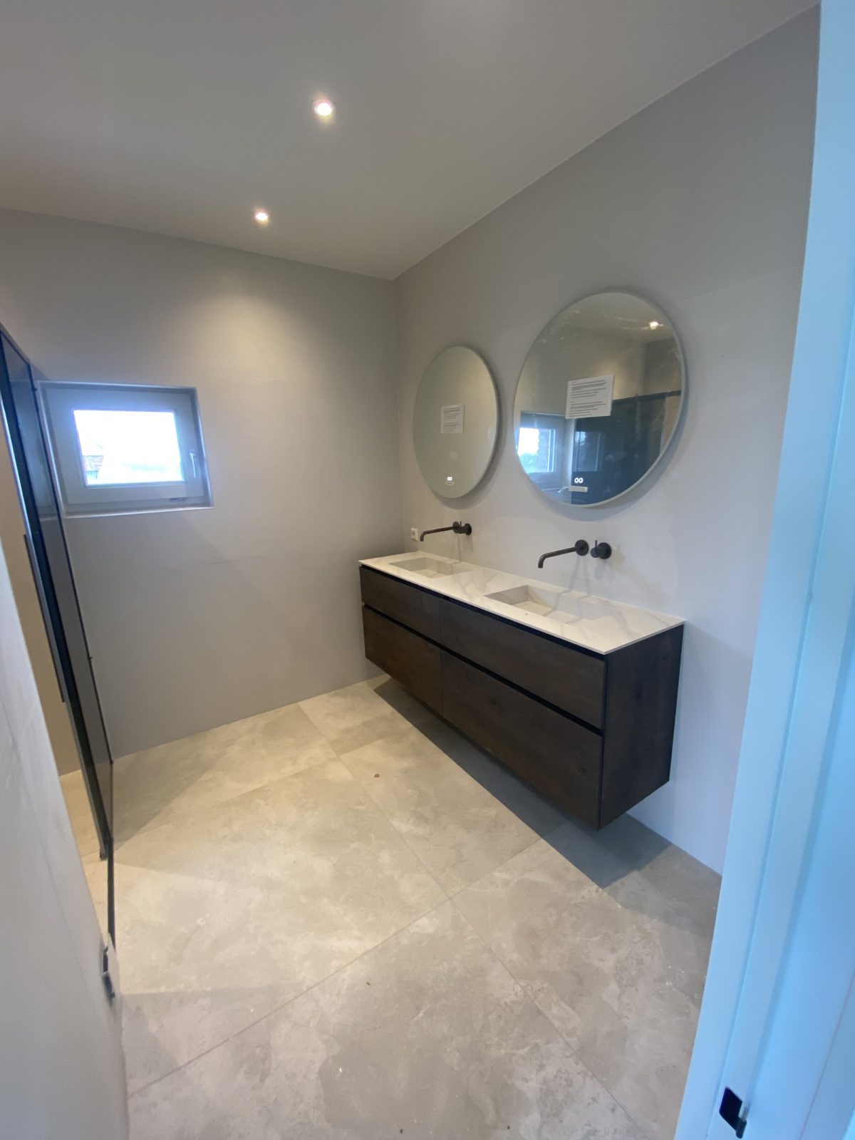 Badkamer met grote lichte vloertegels, badmeubel met dubbele wastafel en twee ronde spiegels