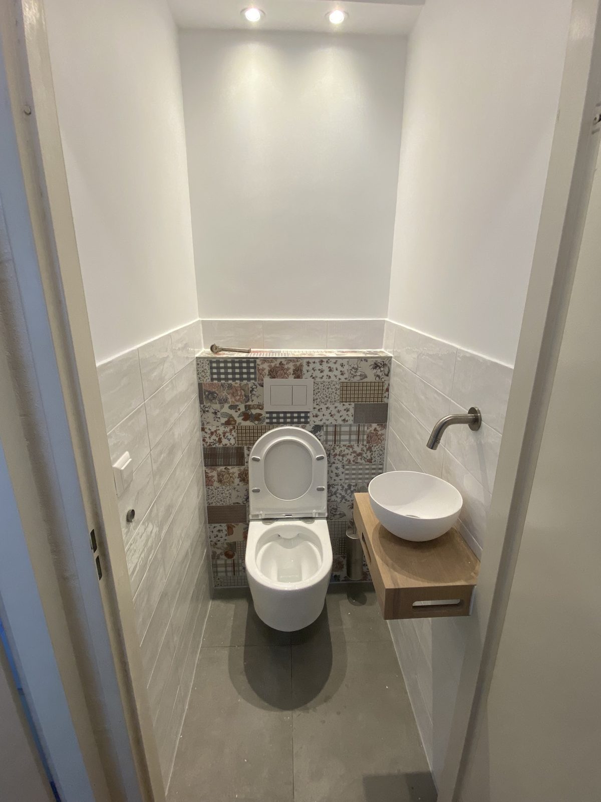 Toilet met een paneel van patchwork-patroontegels achter het hangtoilet en een witte waskom op houten plank