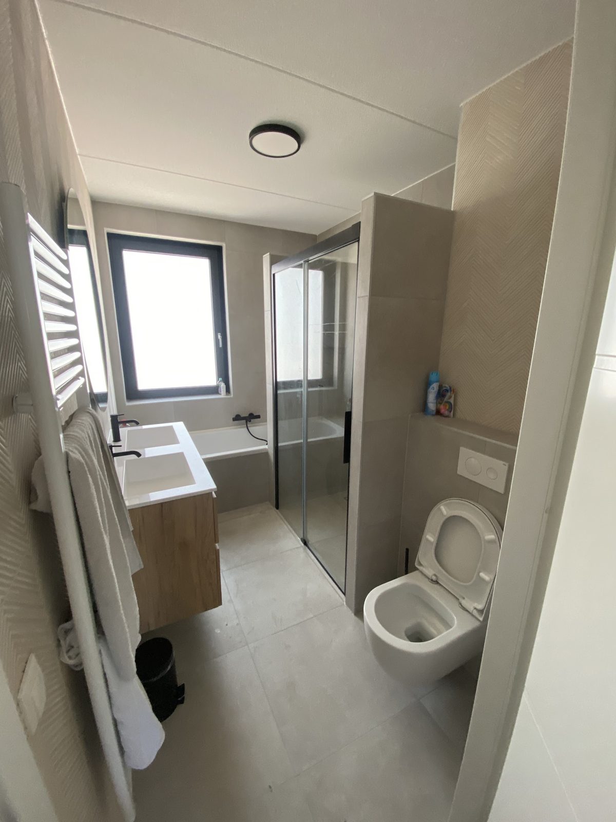 Lichte badkamer met bad achter glas, hangend toilet en houten badmeubel